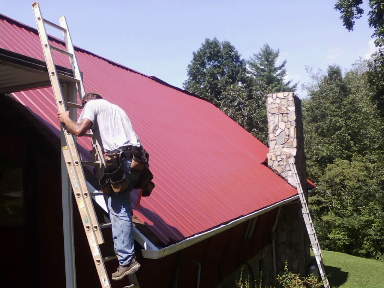 Roofer on a ladder beside a red metal roof and stone chimney — Soffit & Fascia Repair by Roofing Brothers in San Juan Capistrano